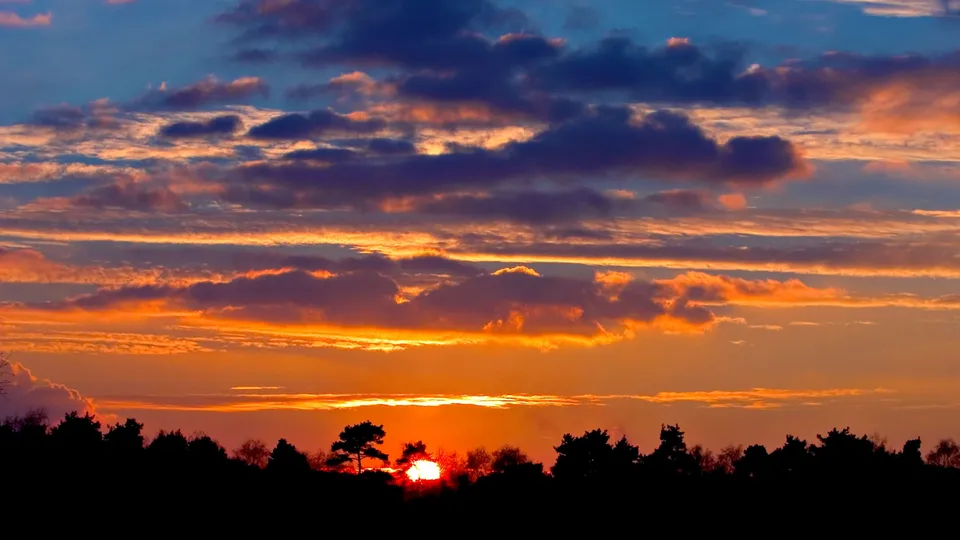 A sunset sky filled with vibrant orange, pink, and purple clouds above silhouetted trees.
