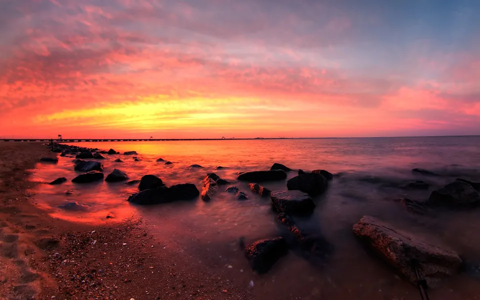 A fiery orange and pink sunset reflects on the calm ocean water, with rocks lining the sandy shore.