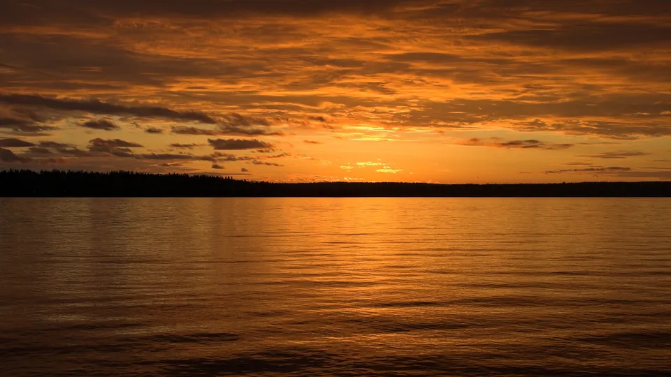 A vibrant orange sky reflects on a calm lake at sunset.