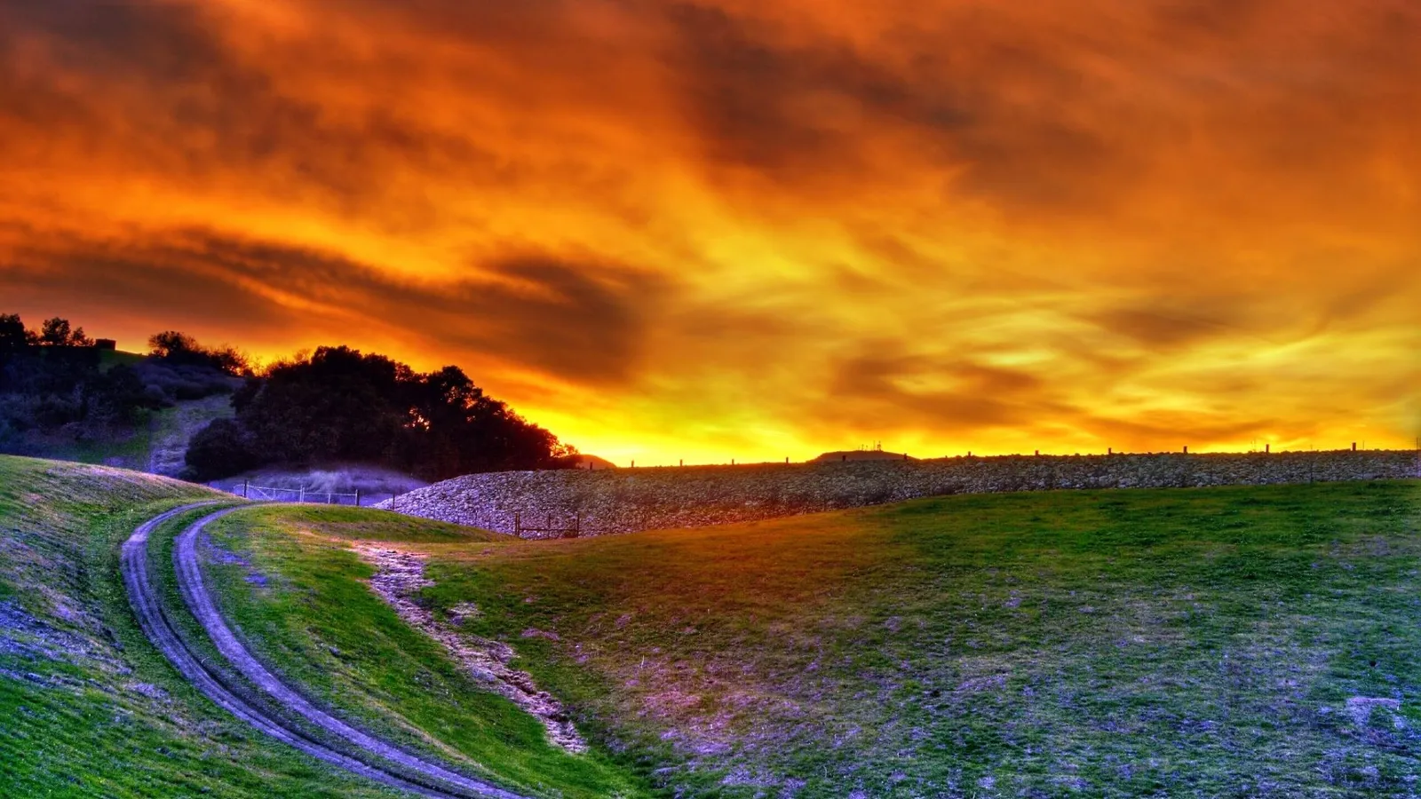 A fiery orange sunset illuminates rolling green hills and a curving dirt path in the foreground.