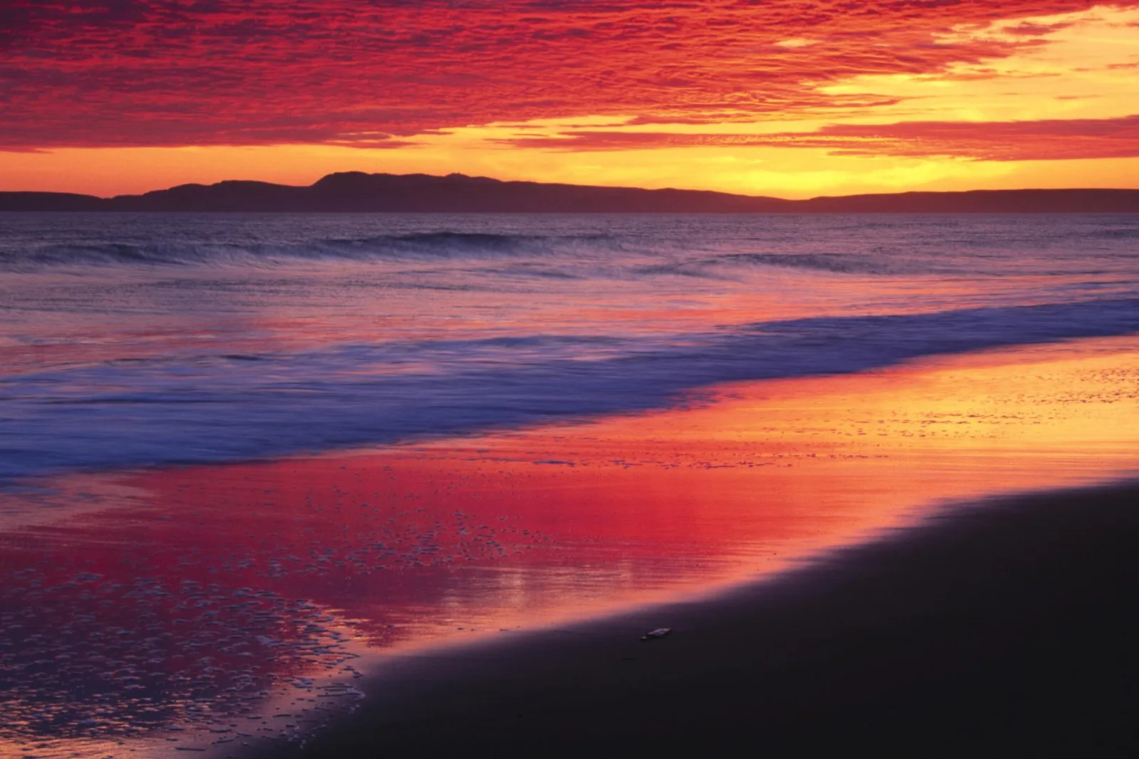A fiery red sunset reflects on the wet sand of a beach, with waves gently rolling in.