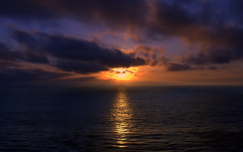 The sun sets over the ocean, with dark clouds illuminated by fiery orange light.