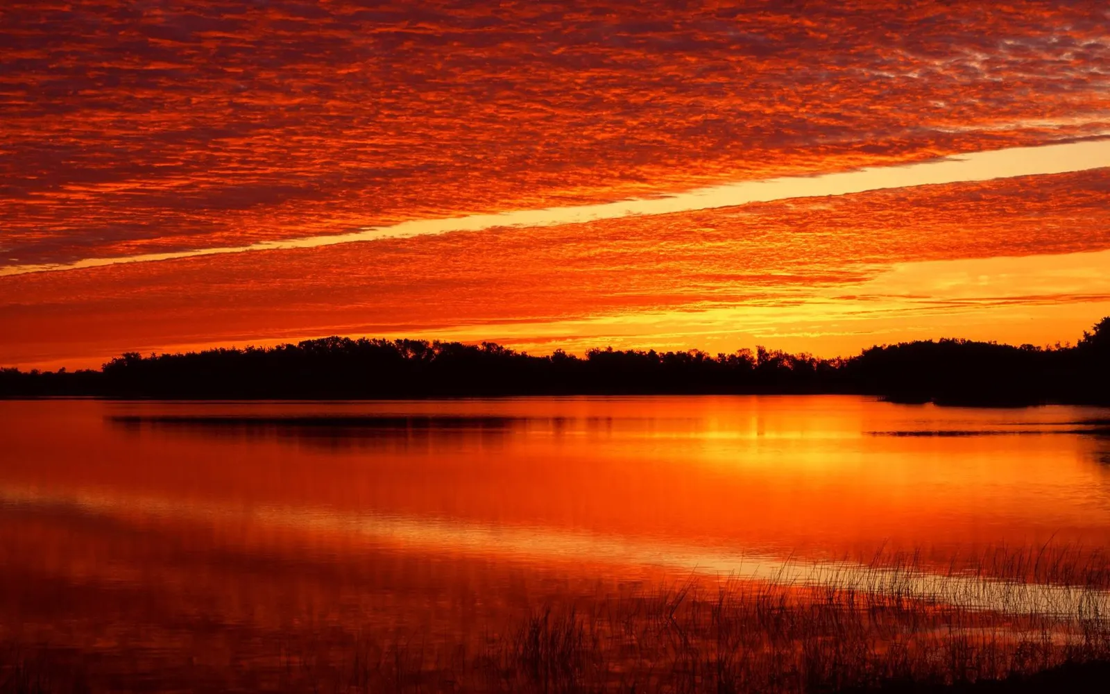 A vibrant orange and red sky reflects on a calm lake, with a silhouetted treeline in the background.