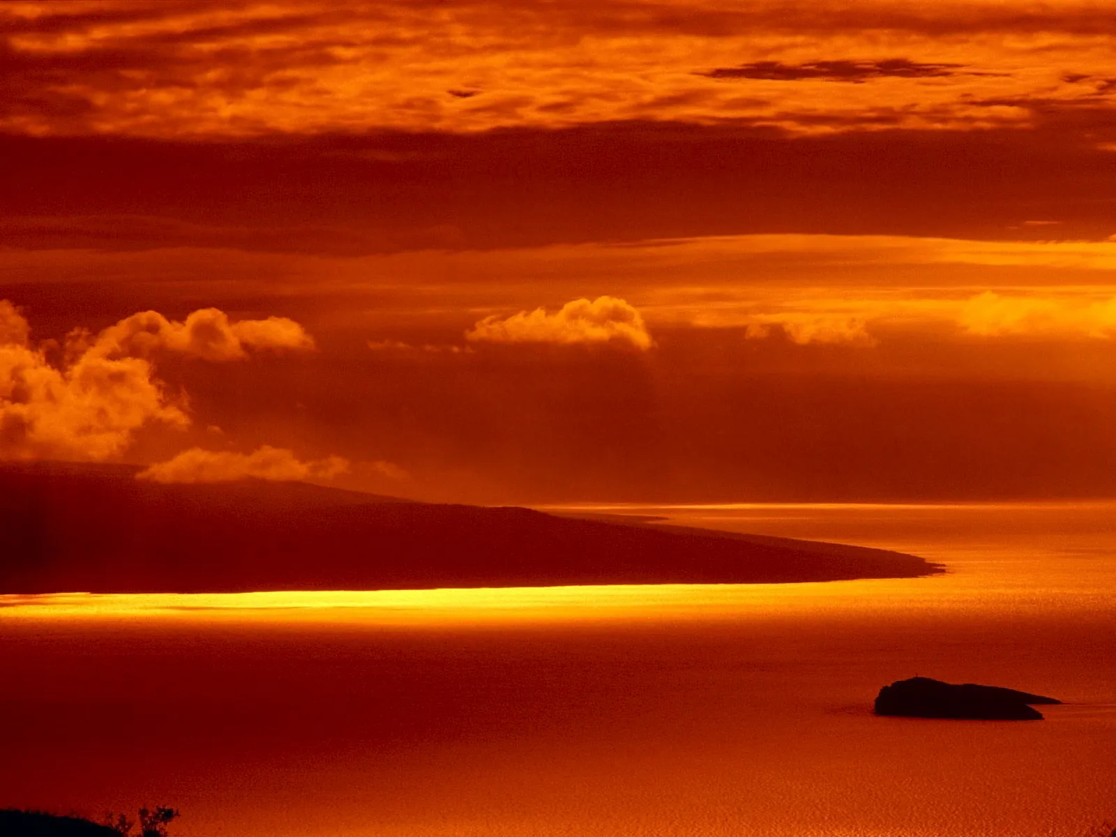 A dramatic sunset with vibrant orange and red clouds over a calm ocean, with a silhouetted island in the distance.