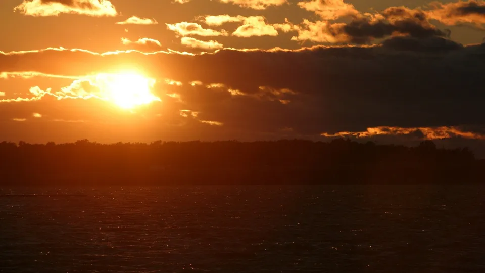 A brilliant orange sun sets over dark ocean waters, partially hidden by dramatic clouds.