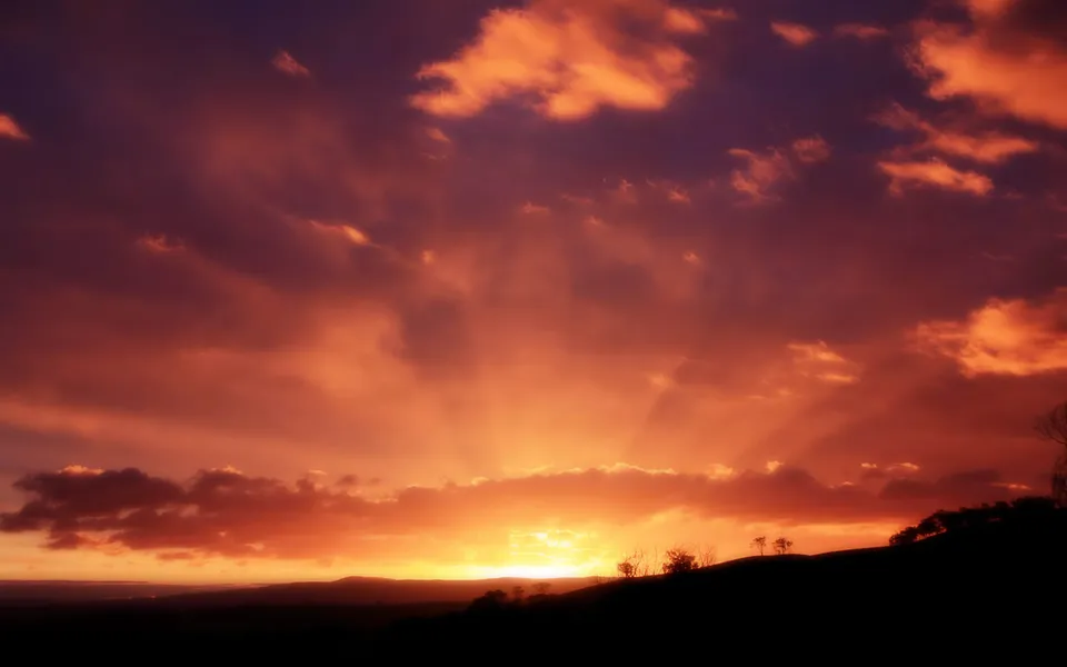 A dramatic sunset with vibrant orange and purple clouds casting rays of light over silhouetted hills.