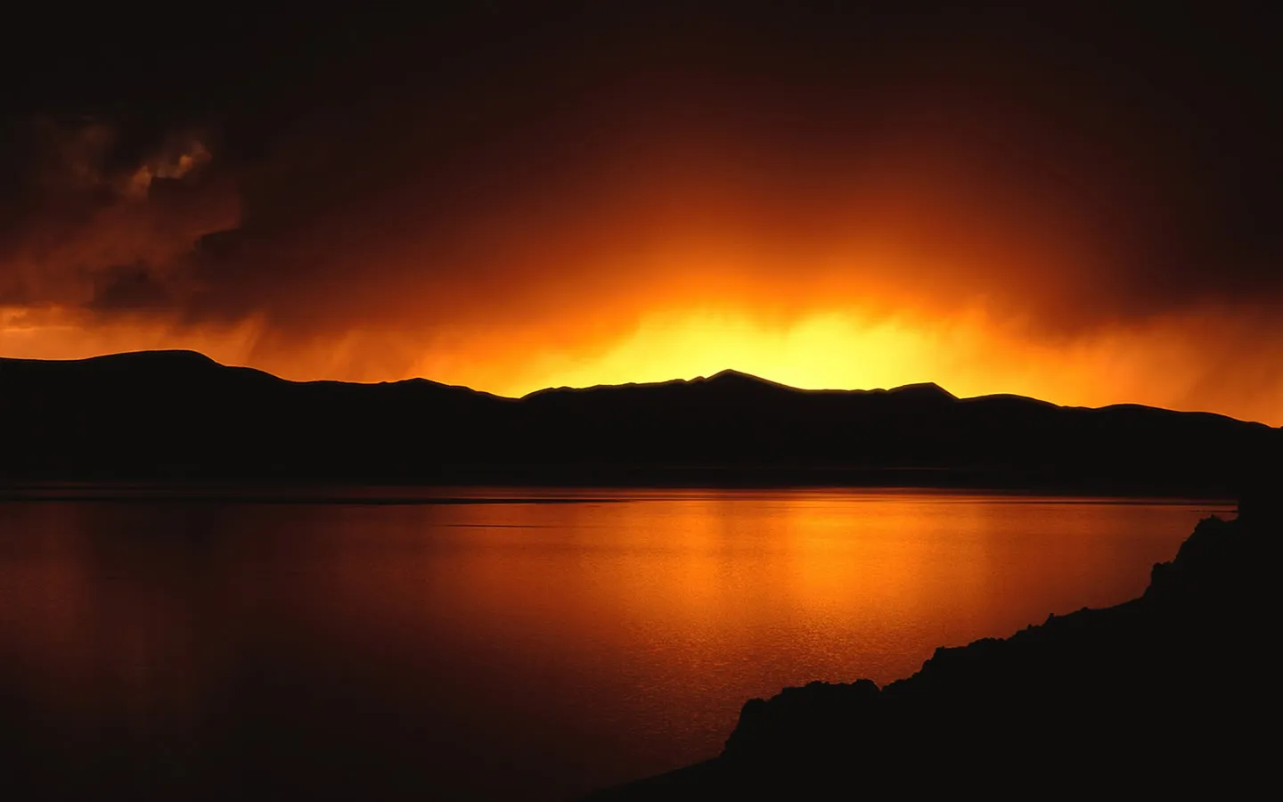 A fiery orange and yellow sunset creates a dramatic sky behind silhouetted mountains and a lake.