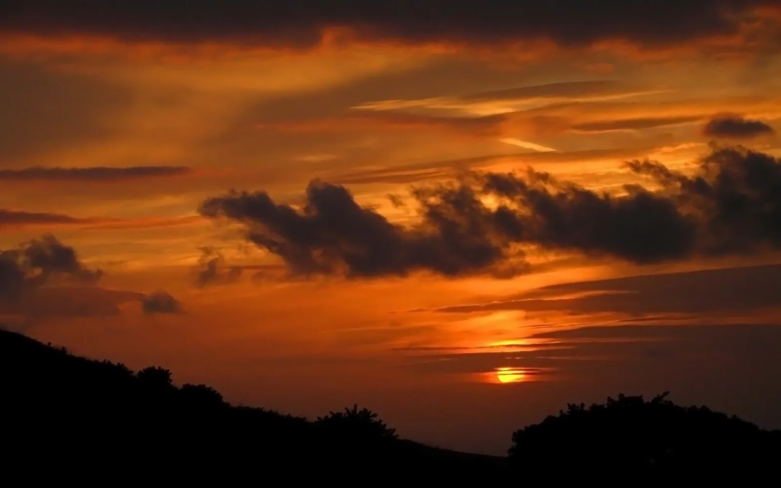 A vibrant sunset with orange and red hues illuminates the sky, with dark clouds and silhouetted trees on a hill.