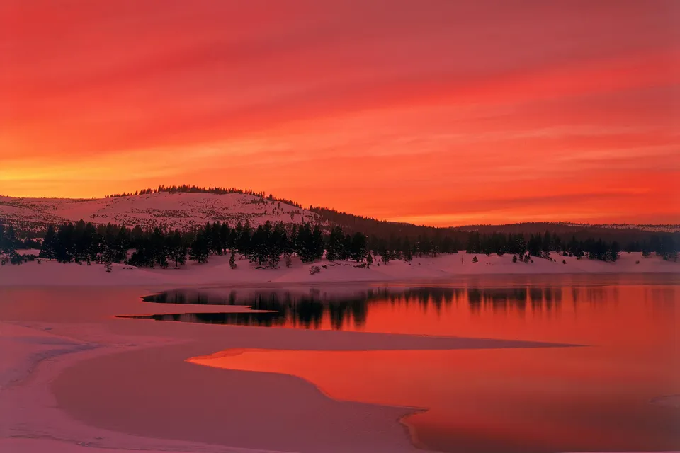 A dramatic sunset with fiery orange and red hues reflects in the calm waters of a snow-covered lake.