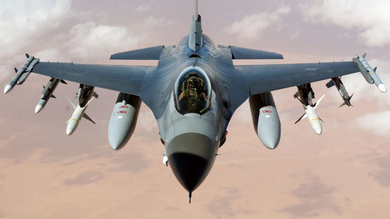 A fighter jet with missiles attached flies through a cloudy sky.