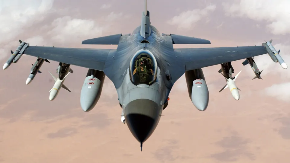 A fighter jet with missiles attached flies through a cloudy sky.