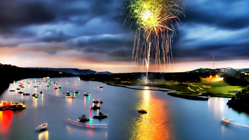 Fireworks explode over a lake filled with boats at dusk, with colorful lights reflecting on the water.