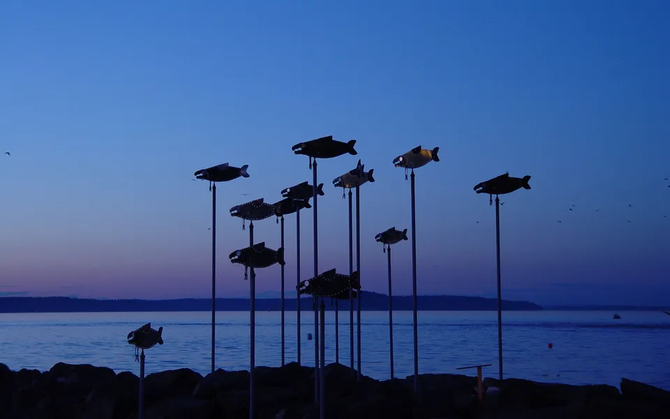 Silhouetted fish sculptures on poles stand against a deep blue and purple twilight sky over a calm body of water.