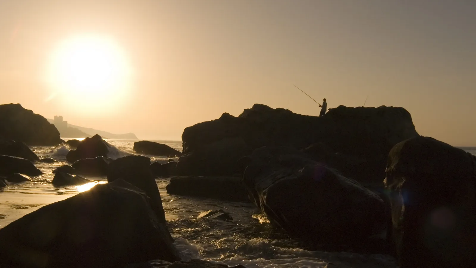 A lone fisherman stands on dark rocks by the sea, silhouetted against a bright, hazy sunset.