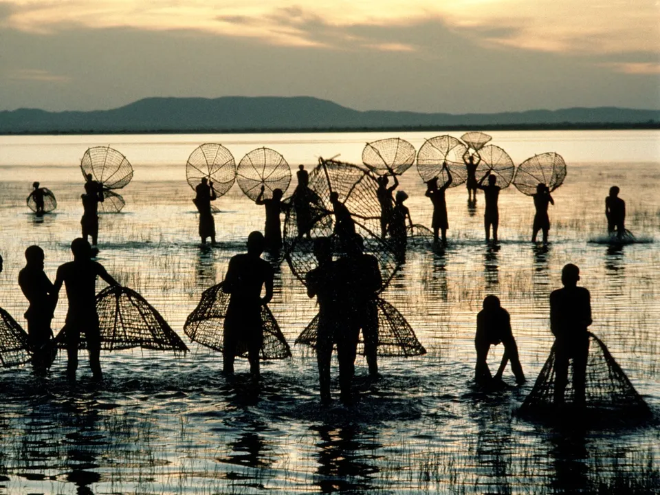 Silhouettes of fishermen wading in shallow water at sunset, holding large nets.