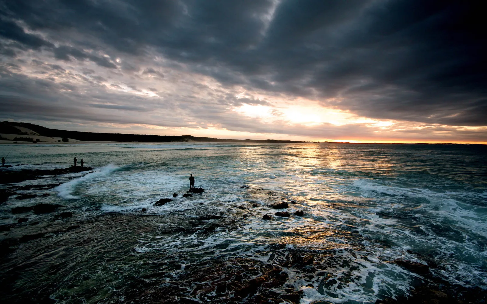 Silhouetted figures of fishermen stand on rocks by the sea as the sun sets.