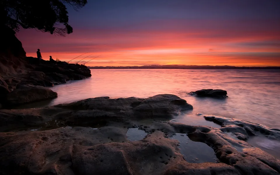 Two people fishing on a rocky shore as the sun sets over the ocean, casting vibrant colors across the sky and water.