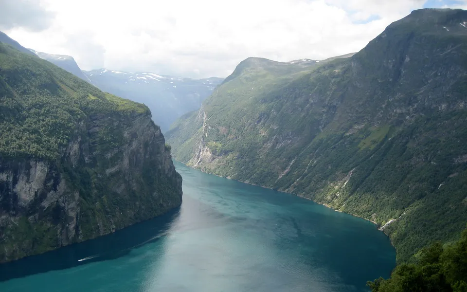 A wide, deep blue fjord is flanked by steep, green, tree-covered mountains under a cloudy sky.