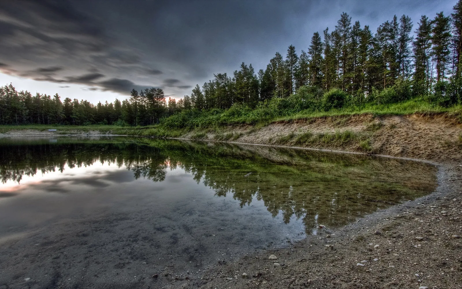 A calm lake reflects a dense forest and a dramatic, cloudy sky at dusk.