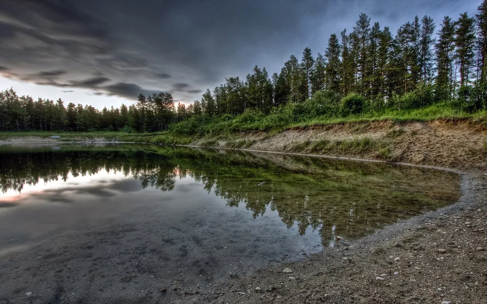 A calm lake reflects a dense forest and a dramatic, cloudy sky at dusk.