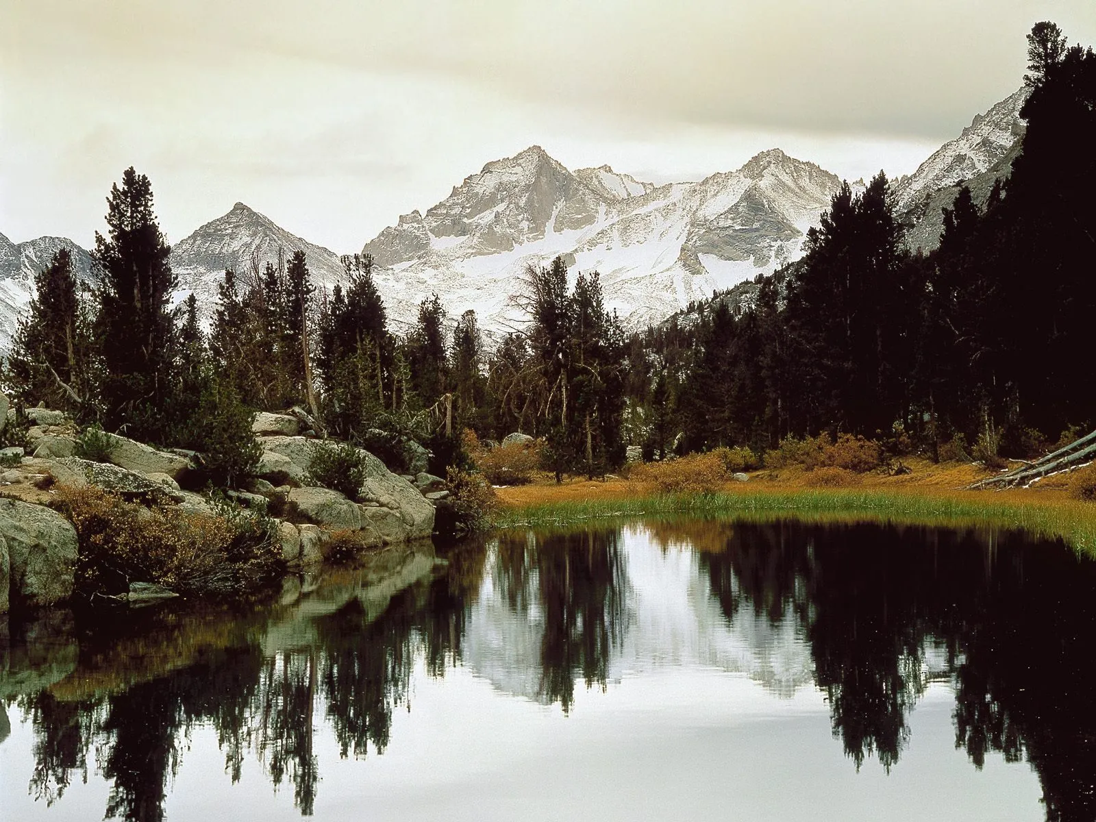 A mountain lake reflects snow-dusted peaks and evergreen trees under an overcast sky.