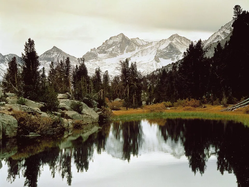 A mountain lake reflects snow-dusted peaks and evergreen trees under an overcast sky.