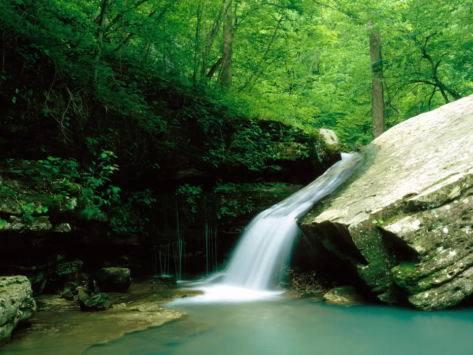 A gentle waterfall flows over large rocks into a clear turquoise pool, surrounded by lush green forest.