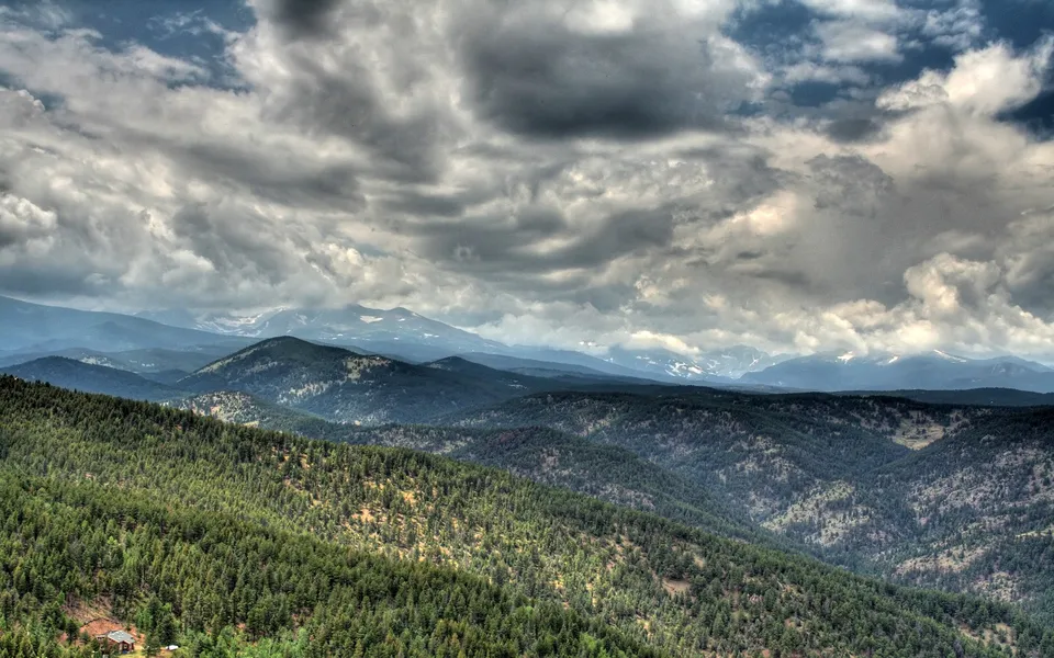 A vast expanse of forested mountains stretches towards distant snow-capped peaks under a dramatic, cloudy sky.