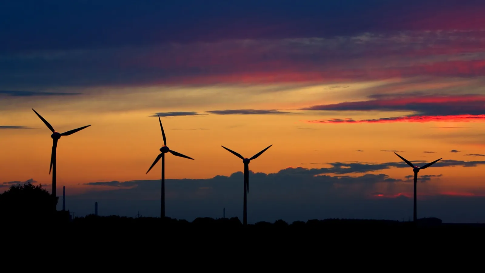 Silhouettes of four wind turbines against a vibrant sunset sky with orange and pink hues.