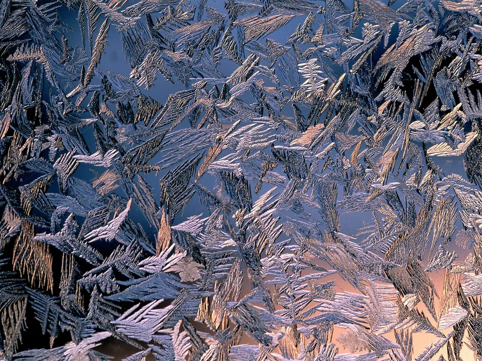 Intricate frost patterns on a window pane with a warm glow behind.