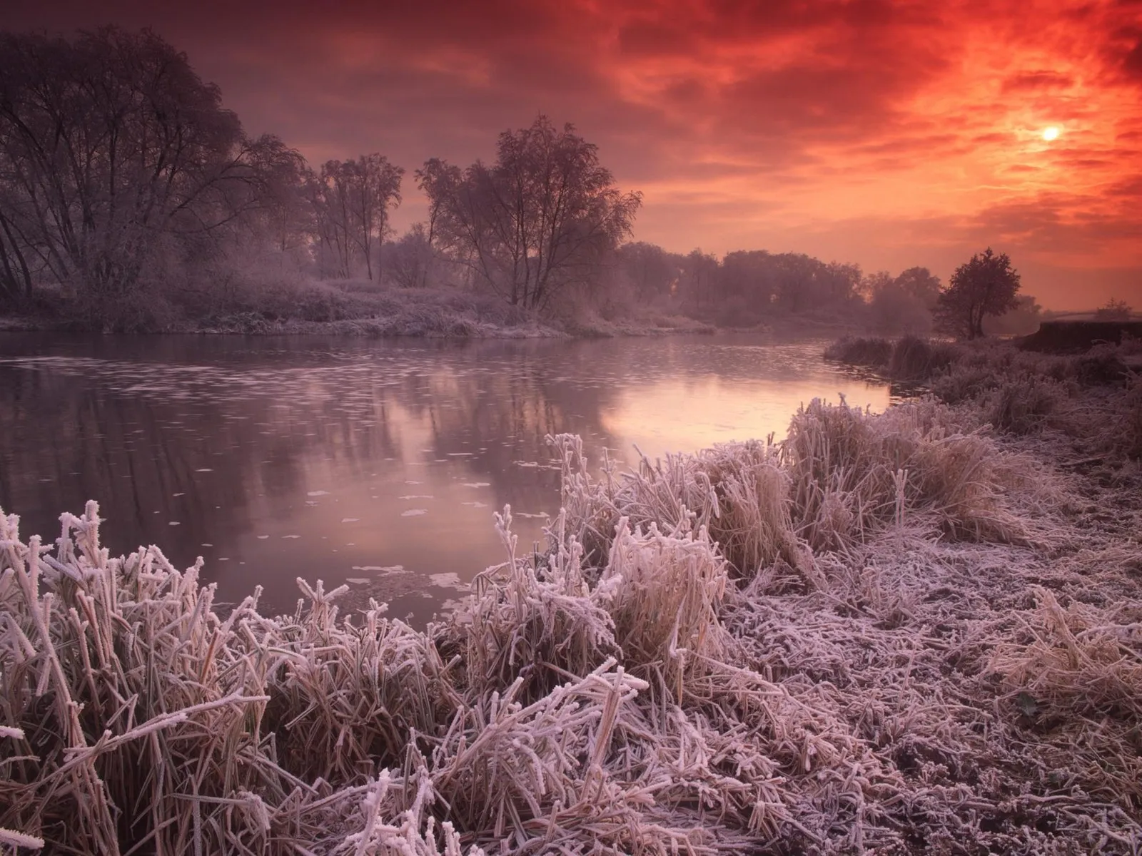 A vibrant sunrise paints the sky with fiery orange and red hues over a frozen river and frost-covered landscape.