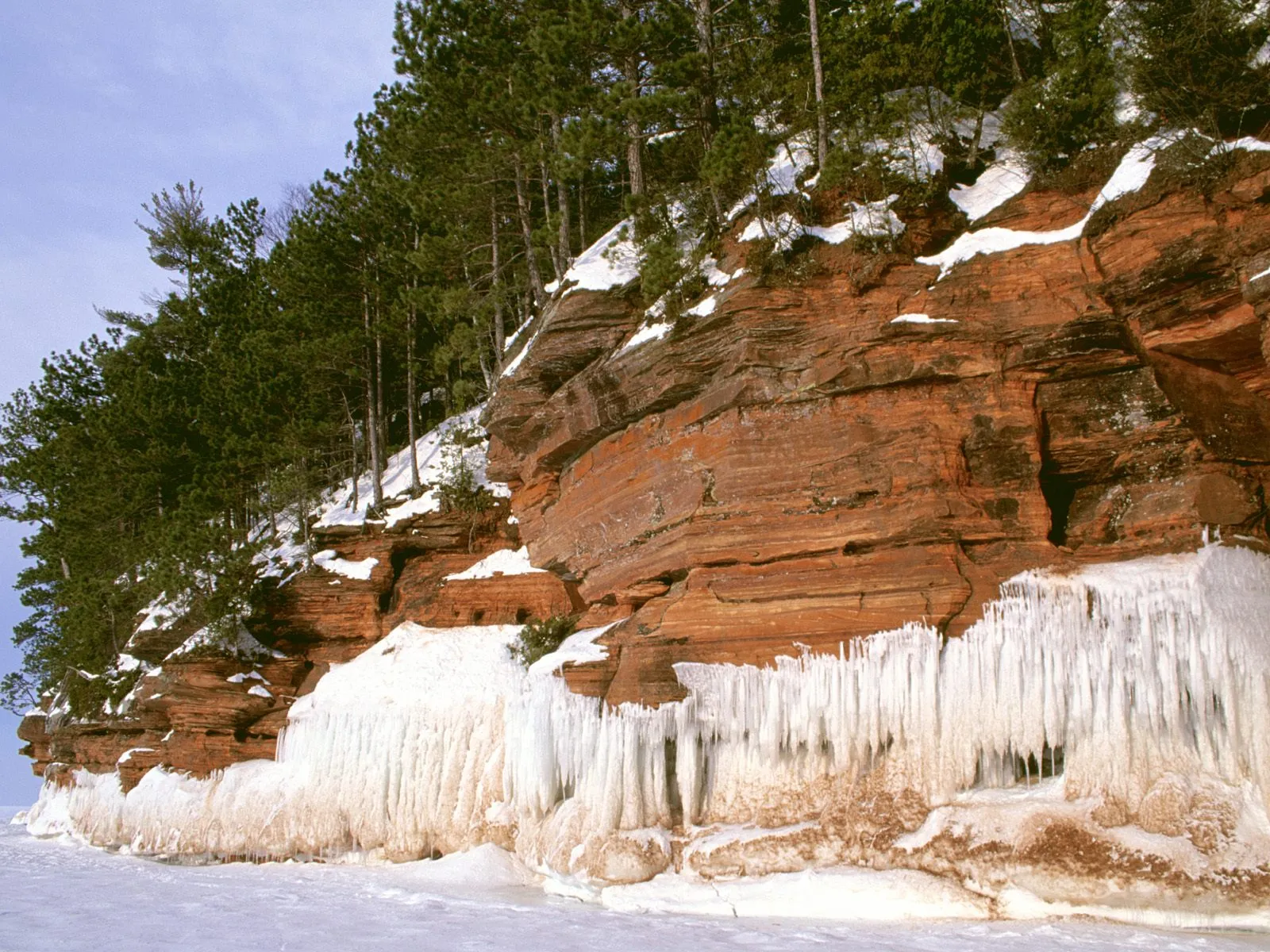 Red sandstone cliffs covered in snow and ice, with pine trees on top, overlooking a frozen lake.