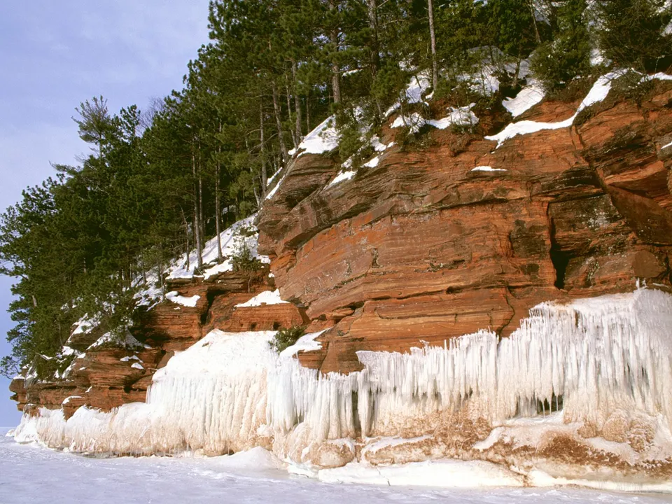 Red sandstone cliffs covered in snow and ice, with pine trees on top, overlooking a frozen lake.