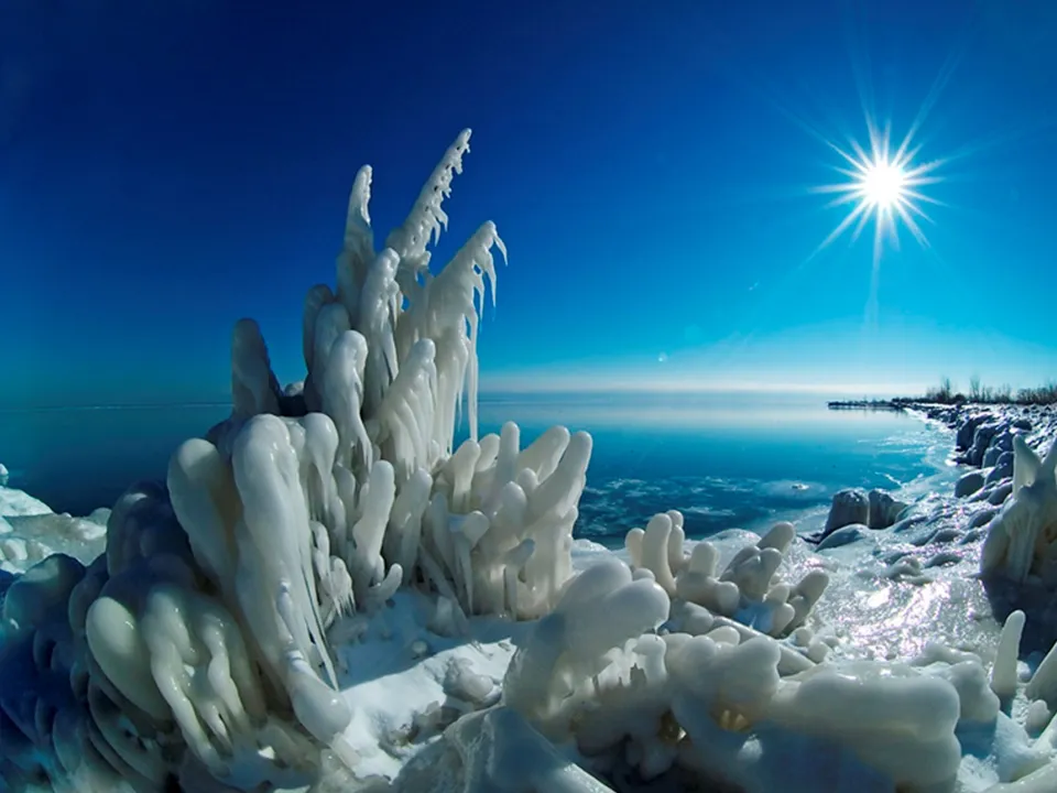 Close-up of intricate ice formations on a snowy shore with a bright sun and calm water in the background.