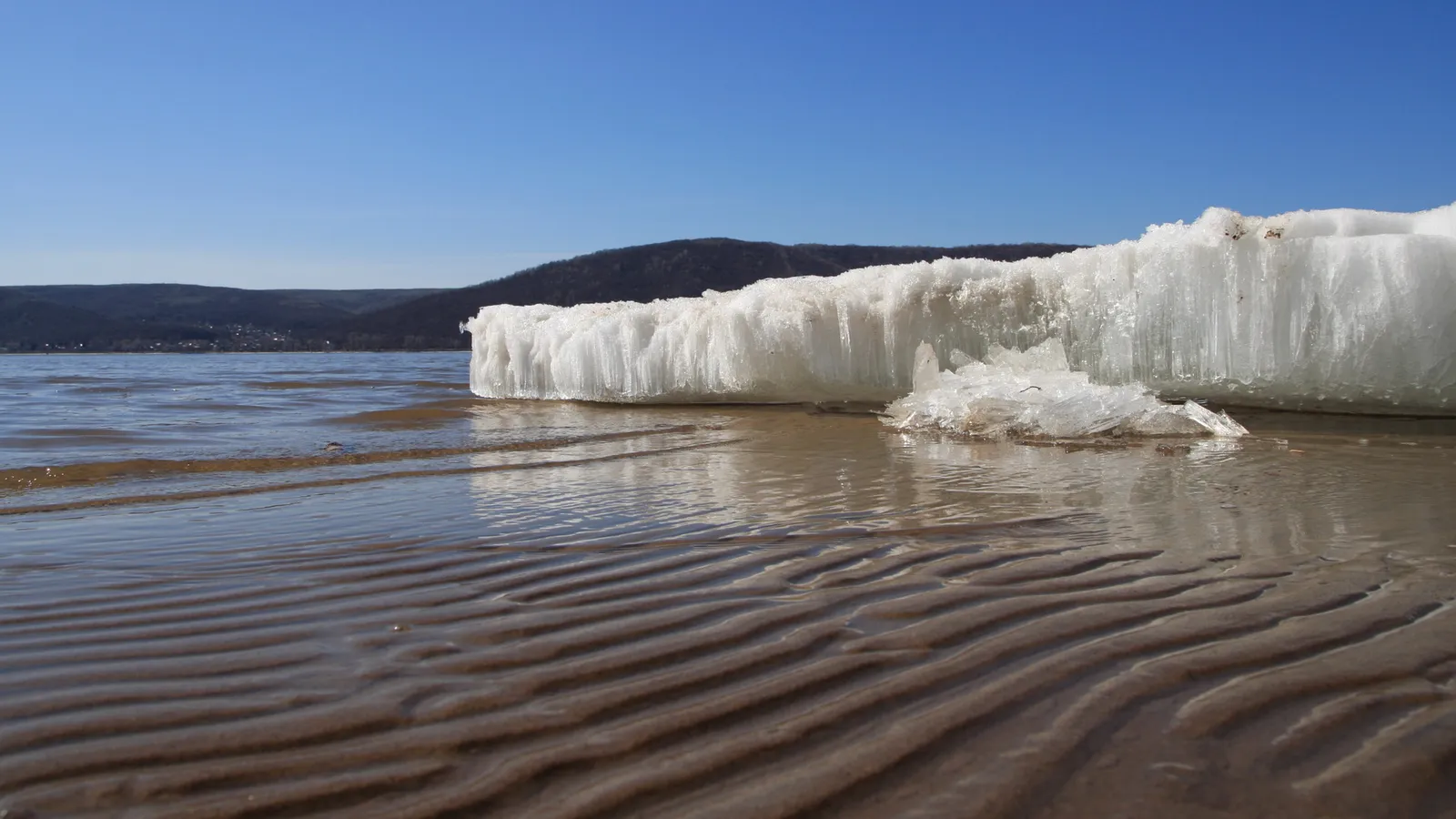 A large formation of ice rests on a sandy riverbank with ripples in the foreground and hills in the background.