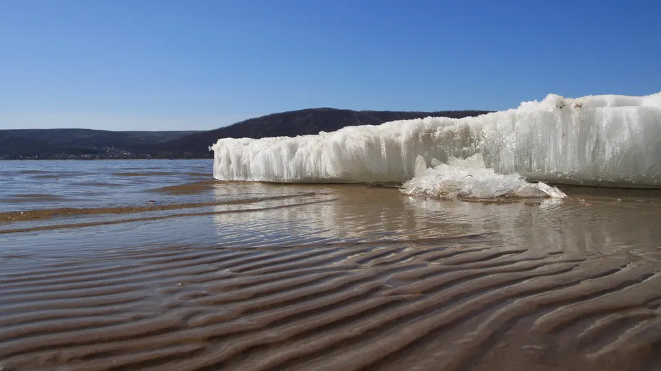 A large formation of ice rests on a sandy riverbank with ripples in the foreground and hills in the background.