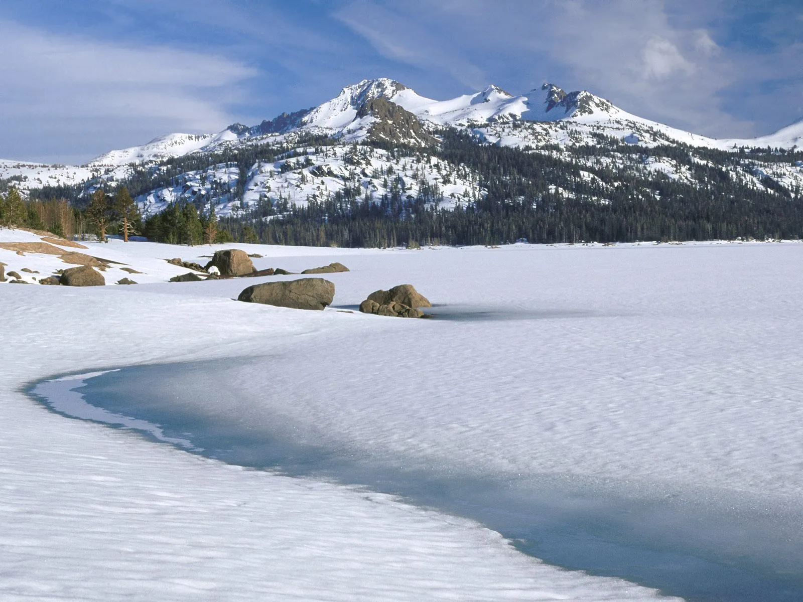 A vast frozen lake with snow-covered mountains and pine trees in the background under a blue sky.