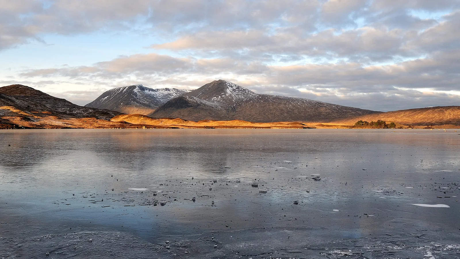 A wide, partially frozen lake reflects the sky, with snow-dusted mountains in the background under a cloudy sky.