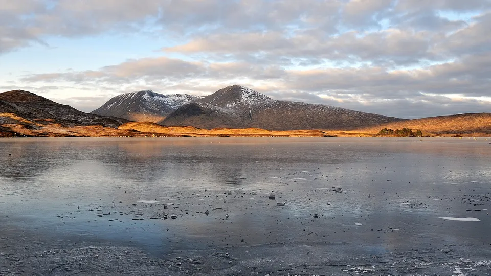A wide, partially frozen lake reflects the sky, with snow-dusted mountains in the background under a cloudy sky.