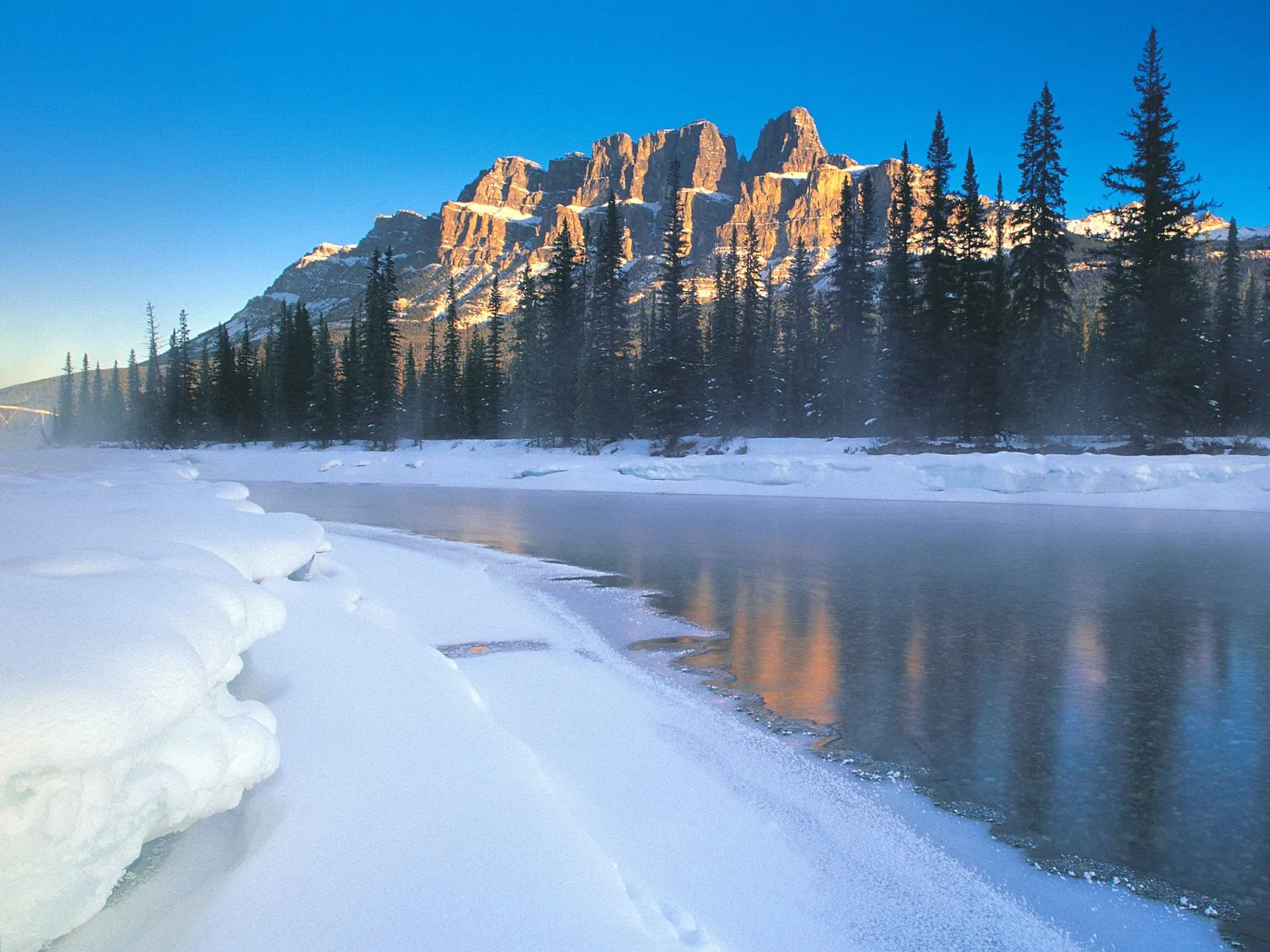 A frozen river flows past snow-covered banks, with pine trees and sunlit mountains in the background under a clear blue sky.