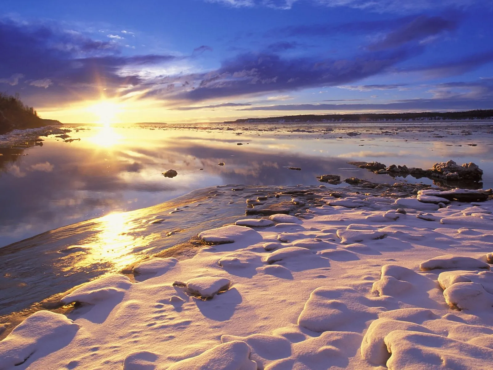 A bright sun rises over a frozen river, casting golden light on snow-covered banks and reflecting in the water.