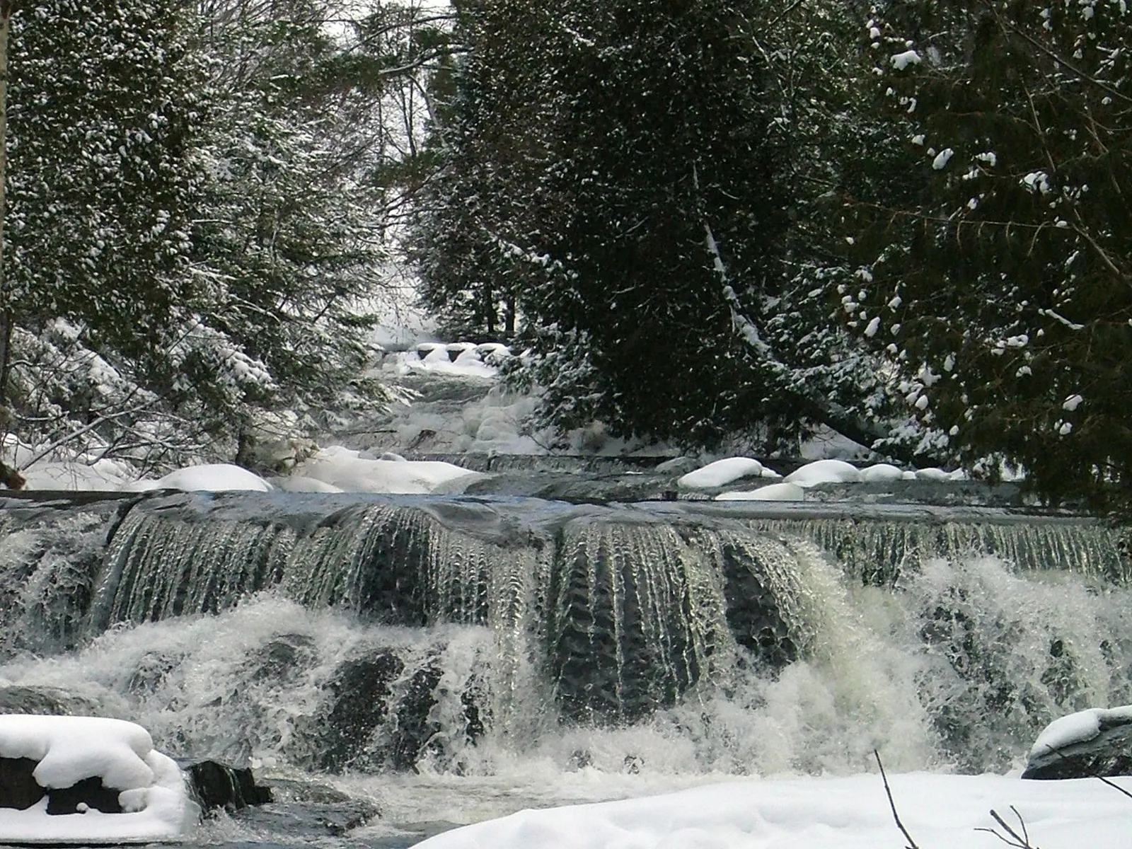 A powerful waterfall flows over dark rocks with snow blanketing the surrounding forest.