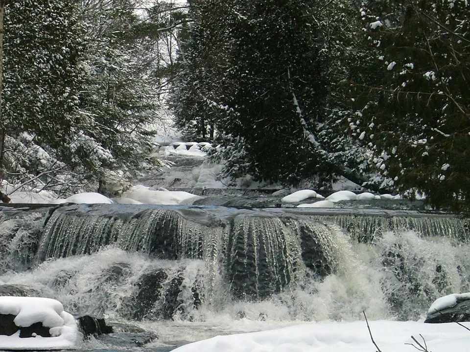 A powerful waterfall flows over dark rocks with snow blanketing the surrounding forest.
