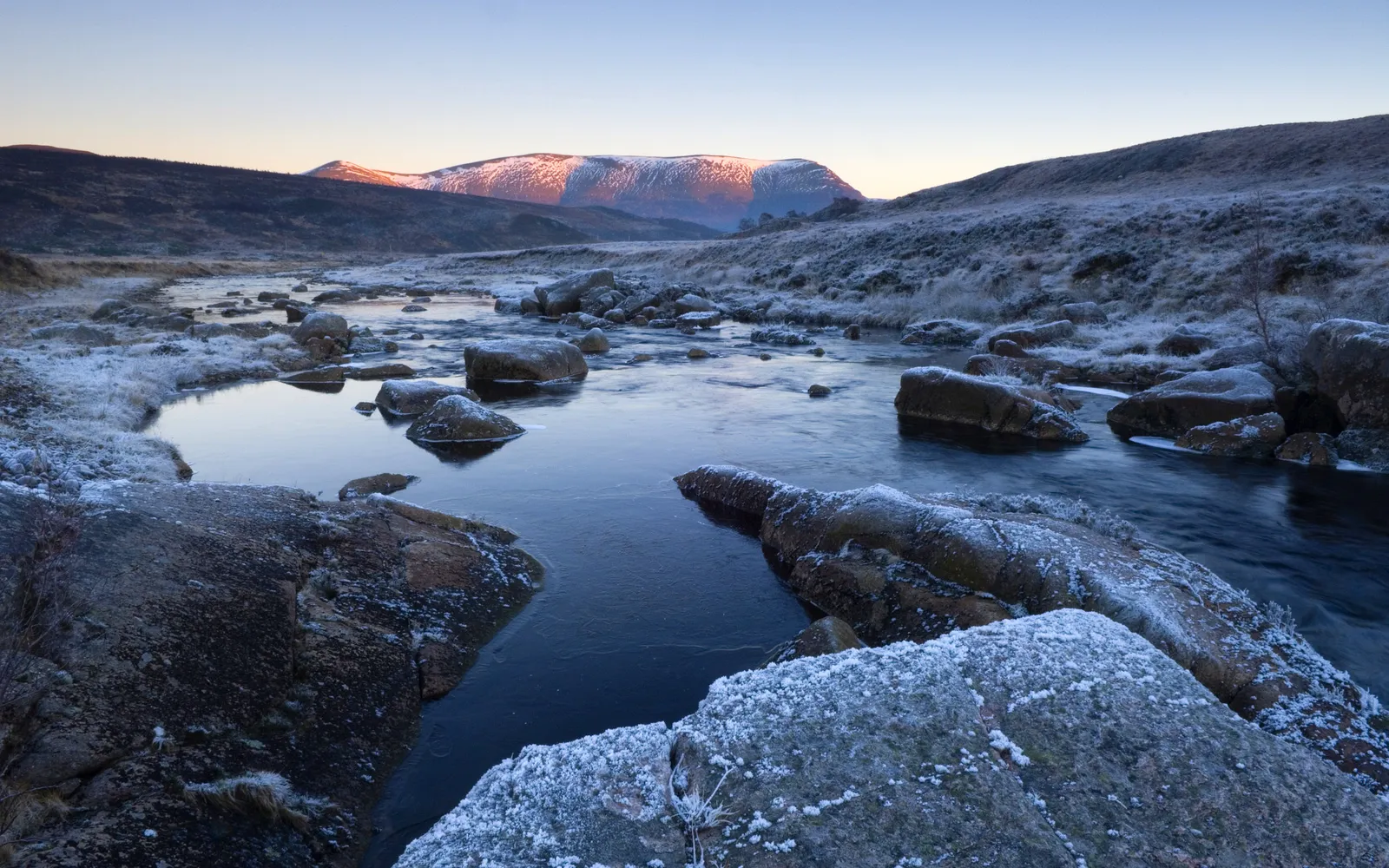 A frozen river flows through a rocky landscape with frost-covered grass and distant mountains.