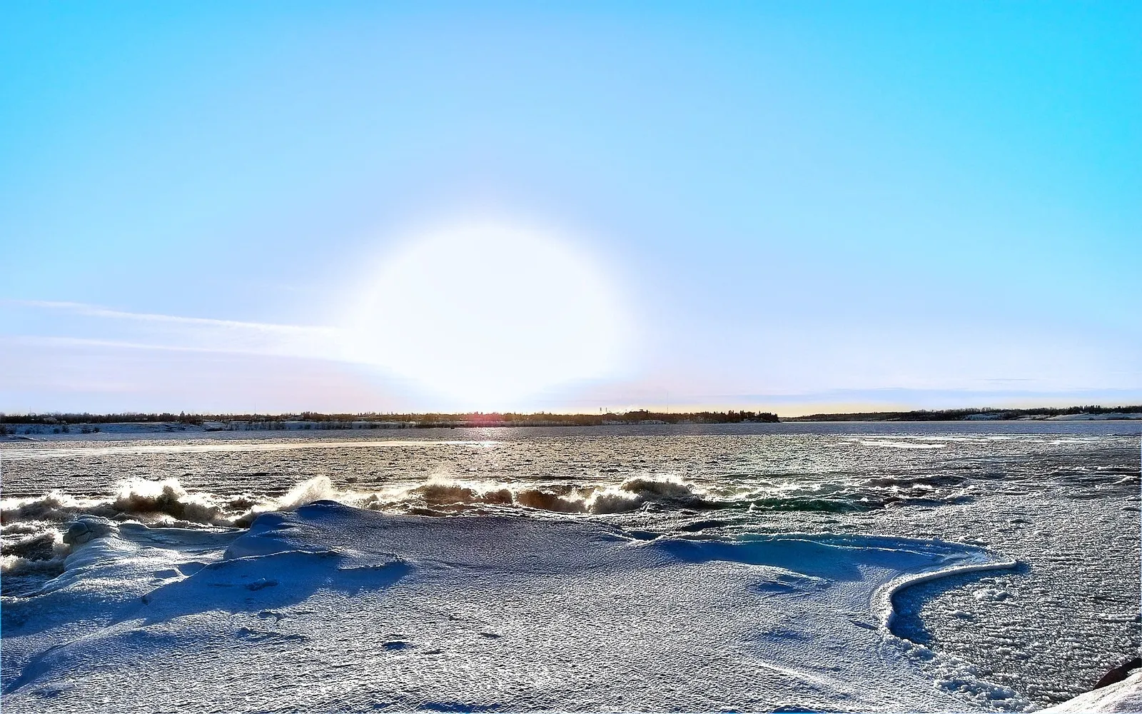 A bright sun shines over a partially frozen river with waves crashing against ice formations.