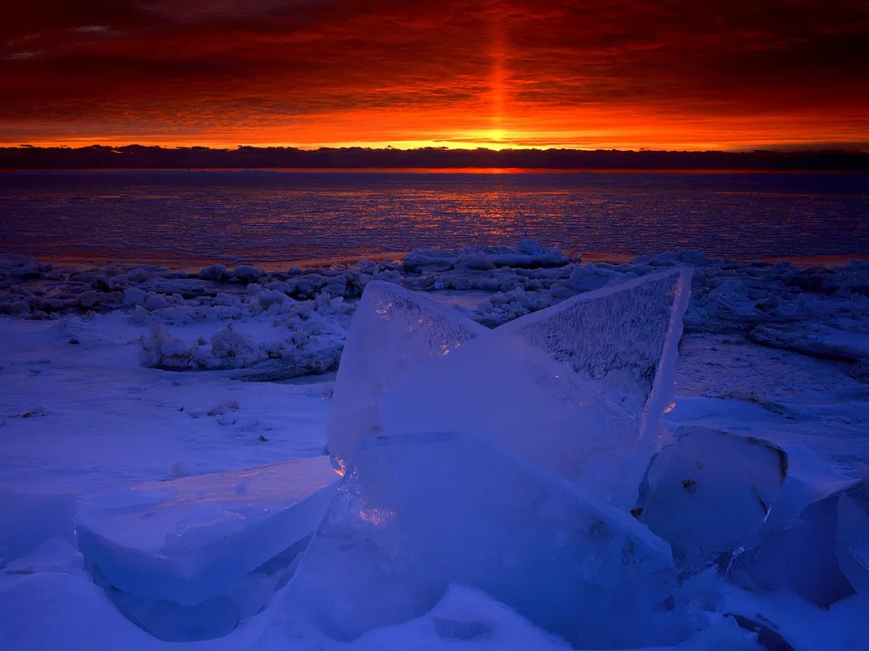 A vibrant sunset with fiery orange and red clouds over a frozen ocean and icy shore.