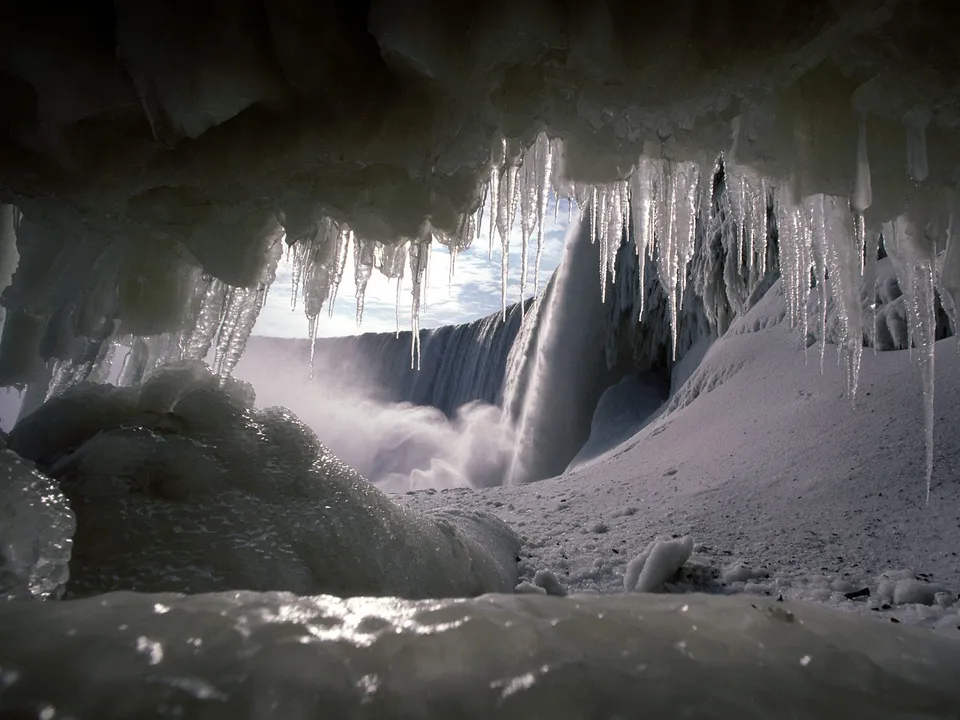 View of a waterfall through a cave covered in ice and icicles, with snow on the ground.