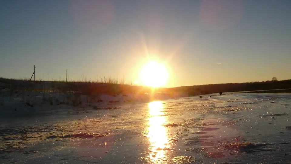 The sun sets over a snow-covered landscape with a frozen body of water reflecting the golden light.