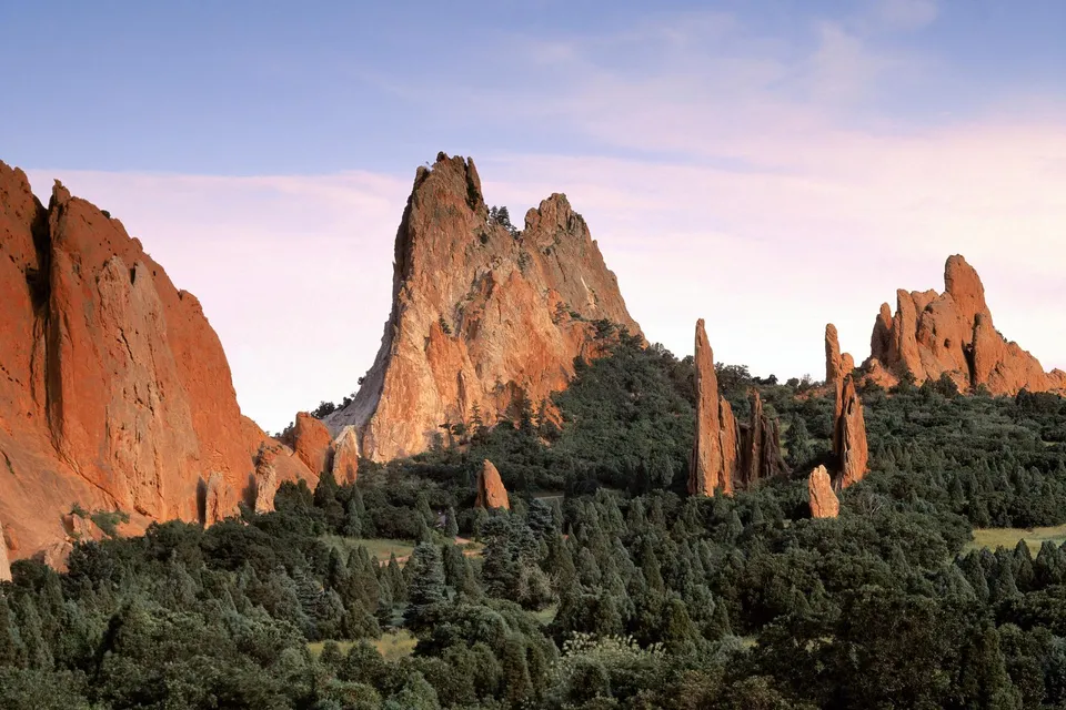 Towering red rock formations rise from a forested landscape under a soft pink and blue sky.