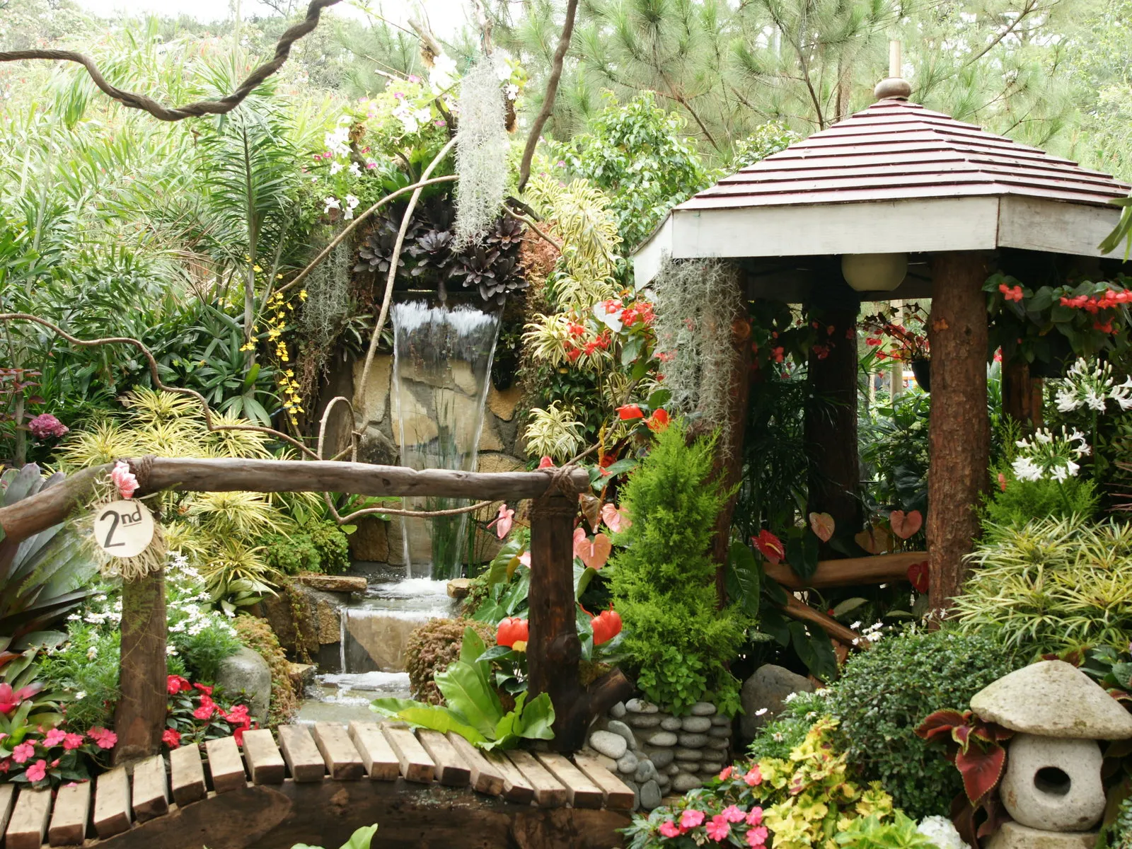 A lush garden scene featuring a small waterfall, a wooden bridge, and a gazebo surrounded by abundant greenery and flowers.